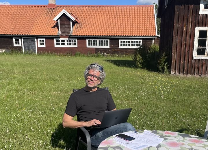 Ola Comstedt, VD på Contrast AB, sitter ute med dator i knät, i ett idylliskt Småländskt landskap bland röda stugor med vita knutar.