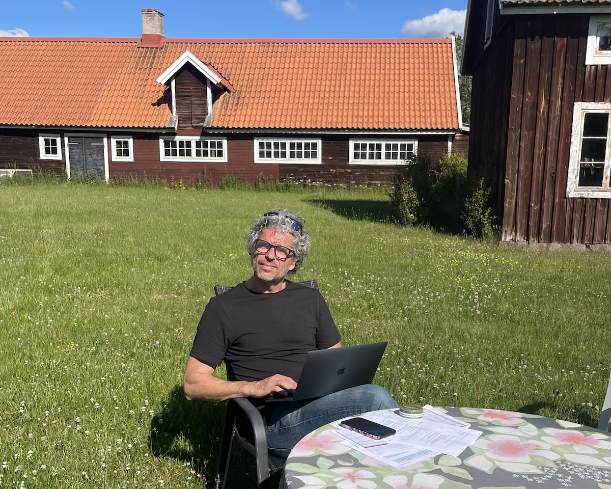 Ola Comstedt, VD på Contrast AB, sitter ute med dator i knät, i ett idylliskt Småländskt landskap bland röda stugor med vita knutar.